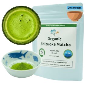 Product photograph featuring Organic Shizuoka Matcha tea showcased in three different forms. The image displays a circular arrangement with the prepared beverage, dry powder, and packaging. On the left side, a dark ceramic bowl contains bright vibrant green matcha tea with a frothy surface displaying small bubbles, demonstrating the traditional whisked preparation. Below it, a small porcelain dish with blue fish and marine motifs contains fine, vivid green matcha powder in its dry form. On the right, a light blue resealable pouch features the product name 'Organic Shizuoka Matcha' prominently displayed on a white label. The package indicates '20 servings' in a blue label at the top corner, with a small illustration of a clay tea bowl. Product details include 'Net Wt. 40g (1.4oz)' and 'Ceremonial Grade' designation. The packaging includes a circular logo featuring a stylized Mt. Fuji with blue water and sky. Descriptive text reads 'Naturally Sweet | Deep Umami Flavor' and 'Premium Cultivar | Stone-milled for Superior Flavor.' A green banner across the top of the label displays the brand's cartoon shark logo with text 'all day i eat like a shark | tea shop.' The composition effectively shows the product's organic certification, premium quality, and traditional Japanese origins, while demonstrating both its powdered form and prepared appearance.