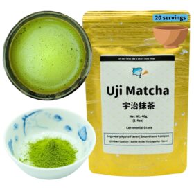 Product photograph of ceremonial grade Uji Matcha tea featuring three elements: a gold metallic package, a prepared matcha drink, and dry matcha powder. On the right, a gold foil resealable pouch displays 'Uji Matcha' in large black text, followed by Japanese characters (宇治抹茶). The package states '20 servings' in a blue label at the top. Product details include 'Net Wt. 40g (1.4oz)' and 'Ceremonial Grade' with descriptive text 'Legendary Kyoto Flavor | Smooth and Complex' and 'Uji Hikari Cultivar | Stone-milled for Superior Flavor.' The packaging features a cartoon shark logo with the text 'all day i eat like a shark | tea shop.' On the left, a dark ceramic bowl contains prepared bright green matcha tea with a frothy surface showing tiny bubbles. Below is a small traditional blue and white porcelain dish containing vibrant green matcha powder. The image showcases both the premium packaging and the product in its dry form and prepared state, highlighting the vivid green color characteristic of high-quality matcha.