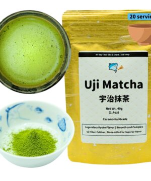 Product photograph of ceremonial grade Uji Matcha tea featuring three elements: a gold metallic package, a prepared matcha drink, and dry matcha powder. On the right, a gold foil resealable pouch displays 'Uji Matcha' in large black text, followed by Japanese characters (宇治抹茶). The package states '20 servings' in a blue label at the top. Product details include 'Net Wt. 40g (1.4oz)' and 'Ceremonial Grade' with descriptive text 'Legendary Kyoto Flavor | Smooth and Complex' and 'Uji Hikari Cultivar | Stone-milled for Superior Flavor.' The packaging features a cartoon shark logo with the text 'all day i eat like a shark | tea shop.' On the left, a dark ceramic bowl contains prepared bright green matcha tea with a frothy surface showing tiny bubbles. Below is a small traditional blue and white porcelain dish containing vibrant green matcha powder. The image showcases both the premium packaging and the product in its dry form and prepared state, highlighting the vivid green color characteristic of high-quality matcha.